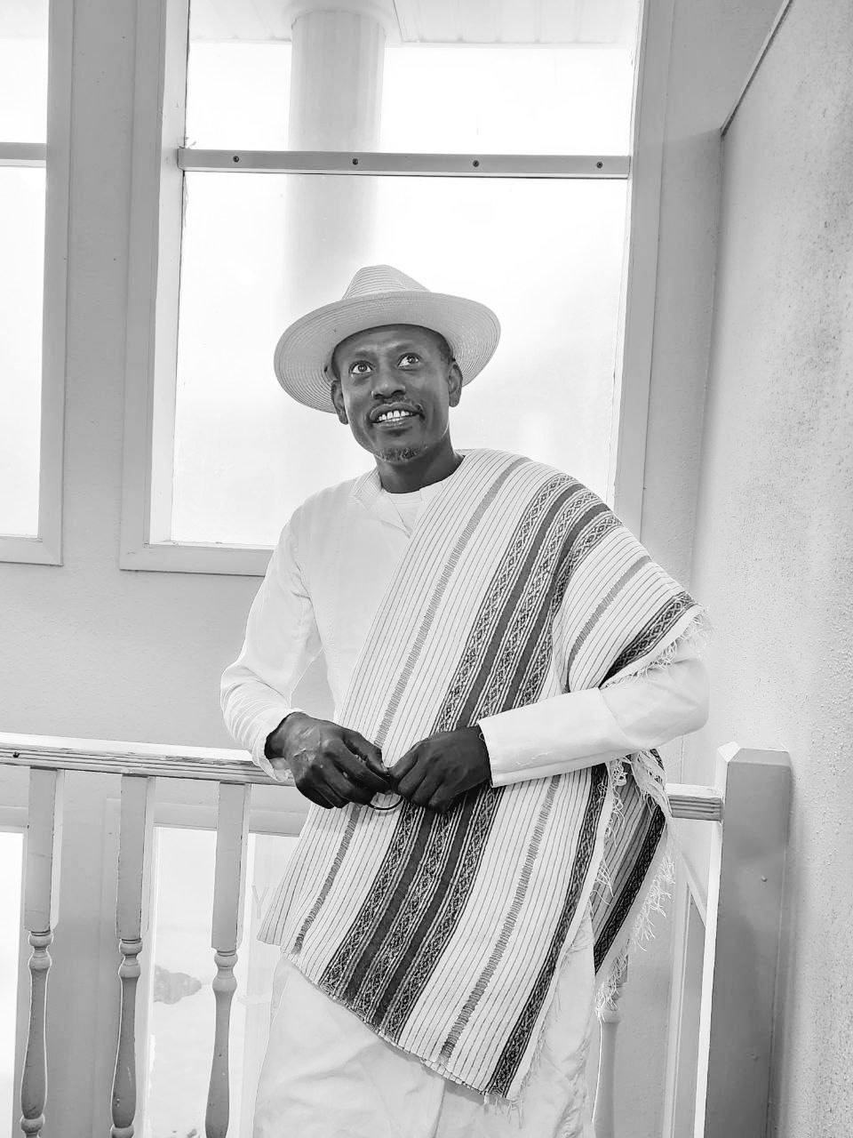 Tariku 80 Shele — portrait in wide-brimmed white fedora, white tunic and woven kuta shawl, holding his glasses
